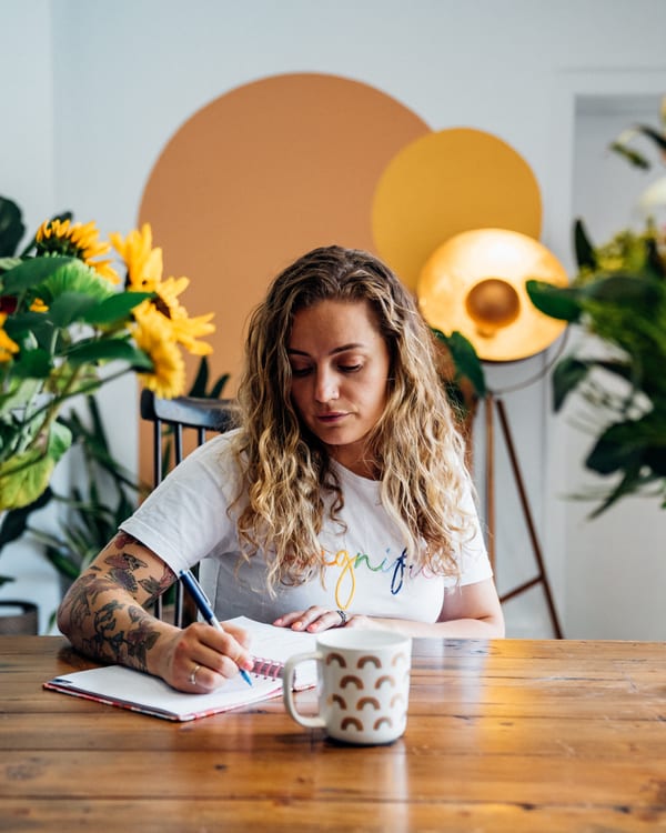 Lashara writing in a notebook at a table, with a coffee and sunflowers beside her